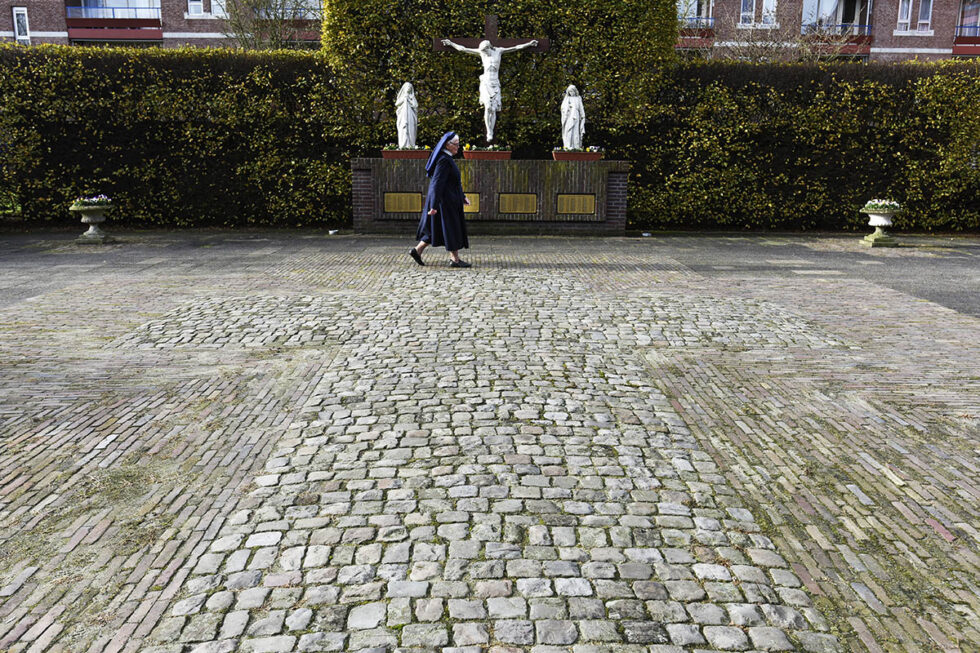2015 Tilburg , Kloostertuin bij het Klooster . Zuster Adelaide van den Berkmortel loopt langs een groot kruis waar o.a. alle zusters van het verleden begraven liggen in een graf met daarop het stenen kruis. Op de achtergrond de namen van de overledenen in de koperen vlakjes onder het kruisbeeld.